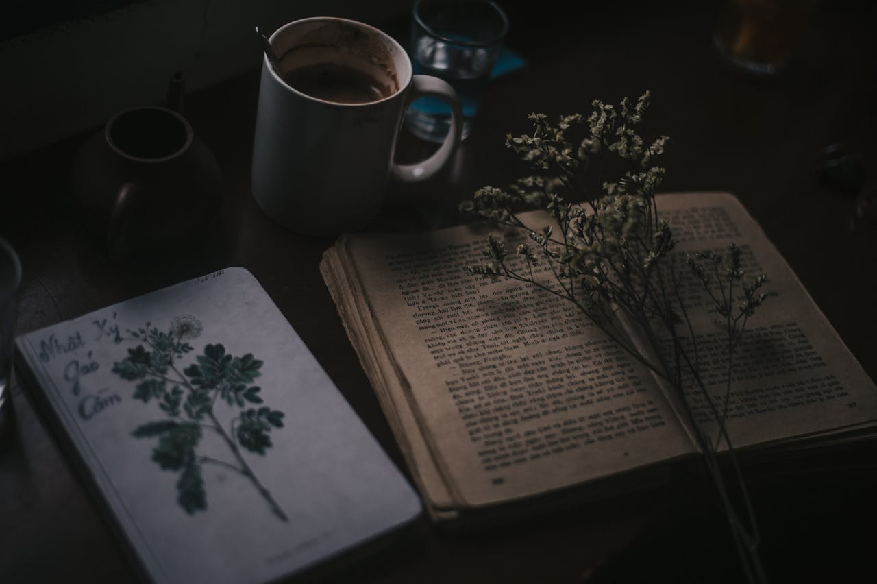 Moody still life of vintage book, coffee mug, and dried flowers on a dark wooden table.