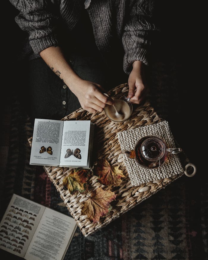 From above of crop faceless person holding cup of fresh coffee while sitting at table and reading book about butterflies